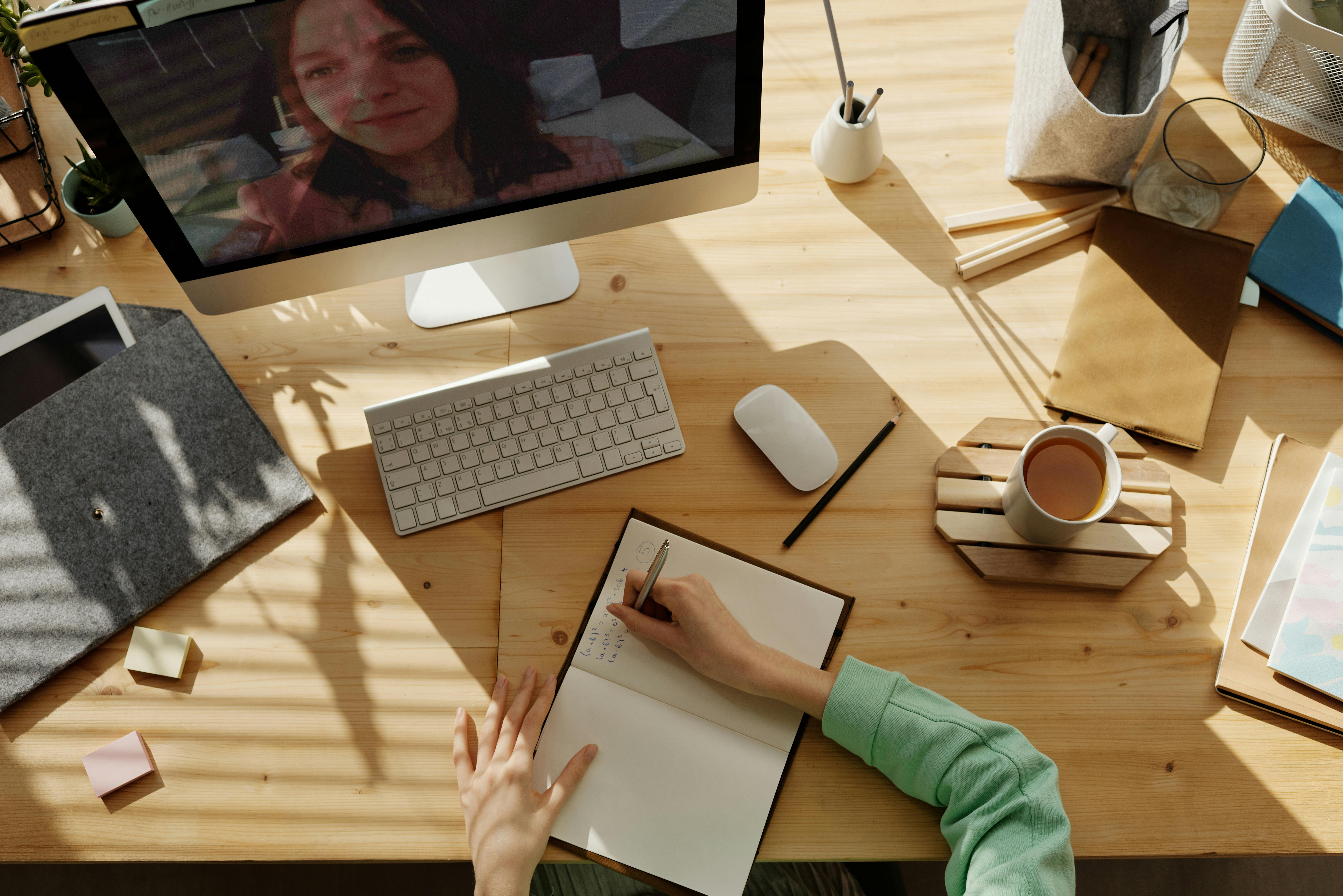 Woman writing in notebook at desk in front of computer with keyboard, mouse, cup of coffee, file envelope, a cup w/pens and pencils.